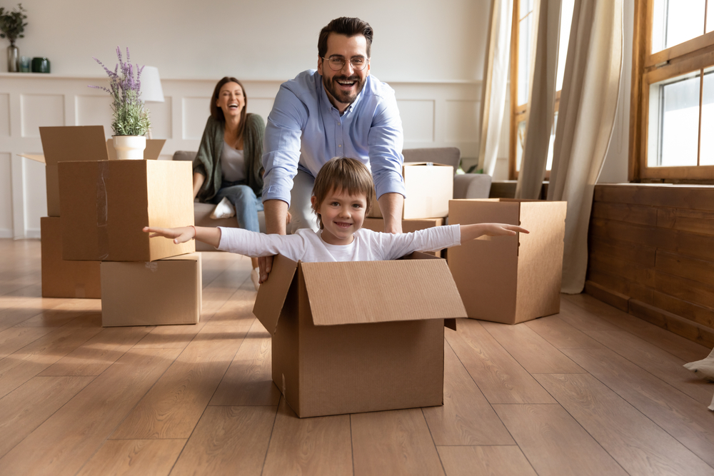 father pushing his kid in a moving box across the room laughing, mother laughing in back
